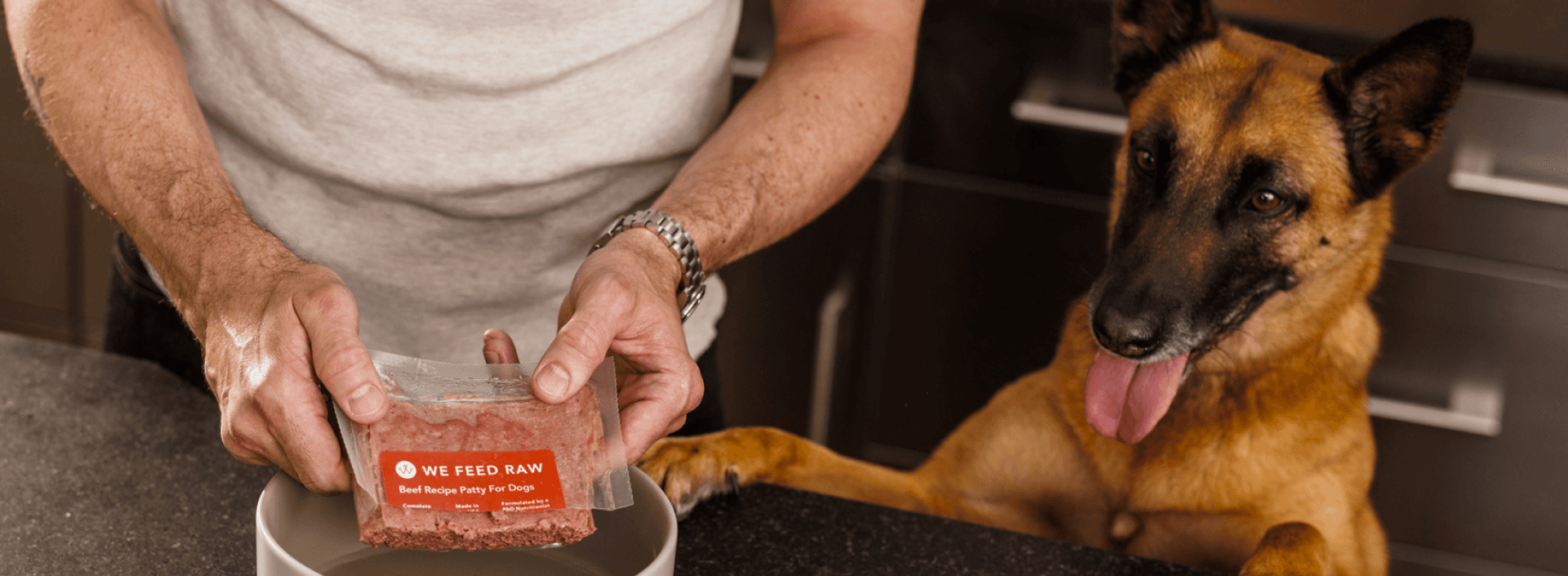 The image shows hands holding a clear package of "We Feed Raw" beef raw dog food over a bowl. A German Shepherd watches eagerly in the background. The raw, red meat is visible through the packaging, emphasizing the product's natural state.