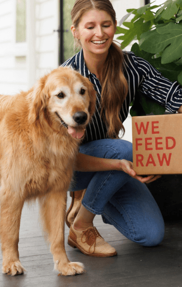 A woman kneeling on her front porch next to her golden retriever dog. She is holding a cardboard box labeled "WE FEED RAW" in red text. The woman is smiling and looking down at the box while the dog stands beside her with its tongue out, looking happy. The background features a potted plant with large green leaves and a black door.