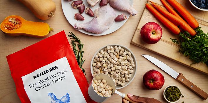 Raw dog food ingredients spread on wooden cutting board including fresh chicken, butternut squash, carrots, apples, blueberries, and a We Feed Raw branded package featuring a blue chicken logo.