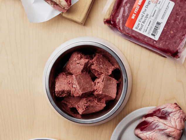 Chunked raw beef dog food in a metal bowl, with We Feed Raw packaging visible in the background